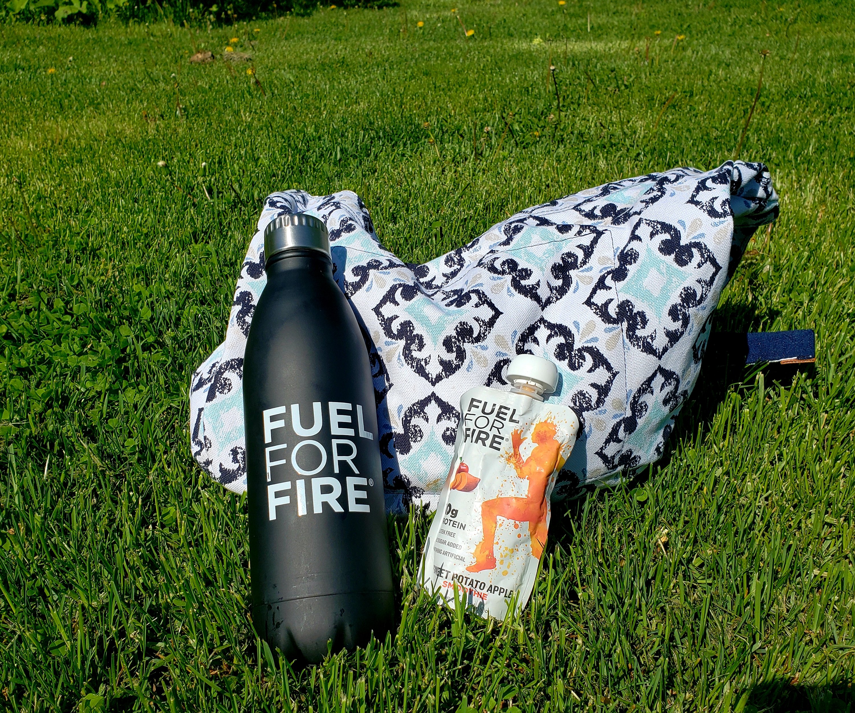 black water bottle with "Fuel for Fire" on it, a sweet potato apple snack pouch and blue patterned bag laying on the grass