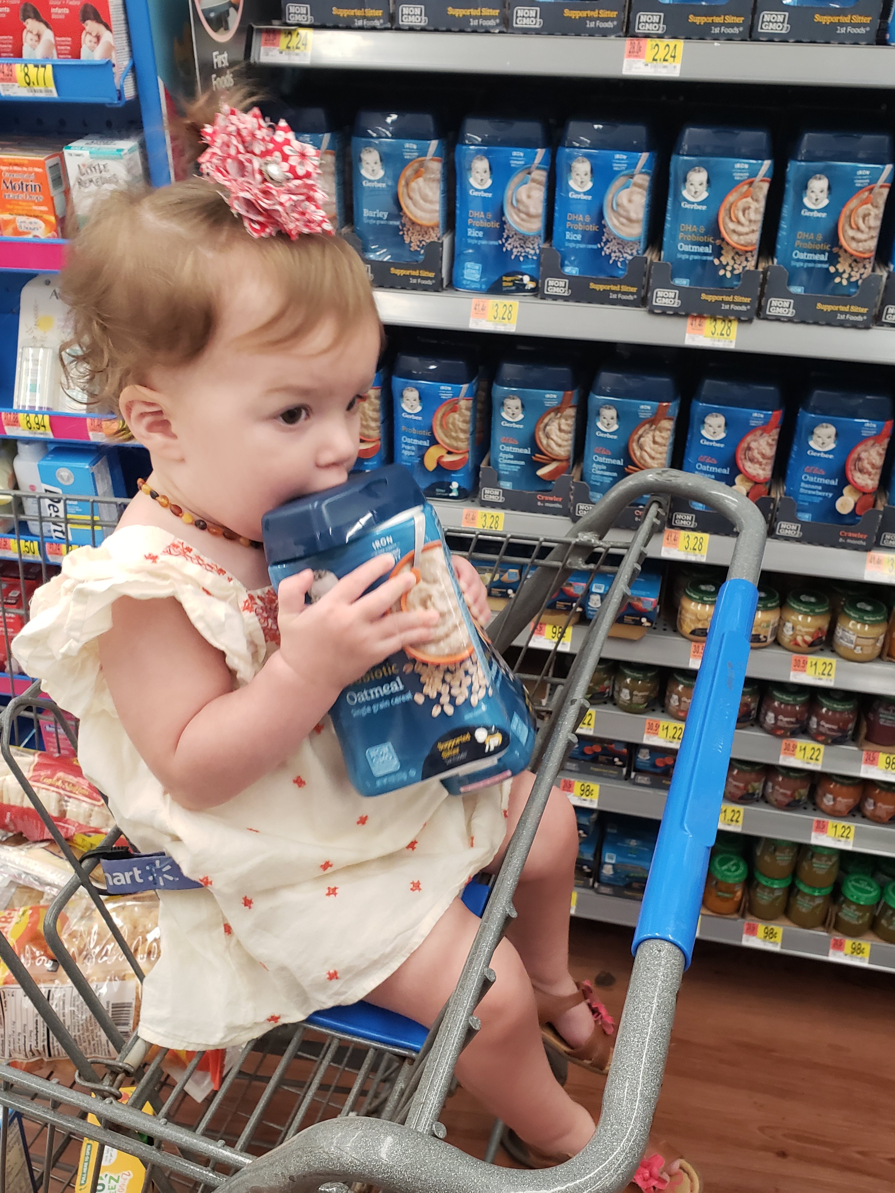 baby girl sitting in a shopping cart in front of Gerber baby food, chewing on a container of Gerber baby ceral