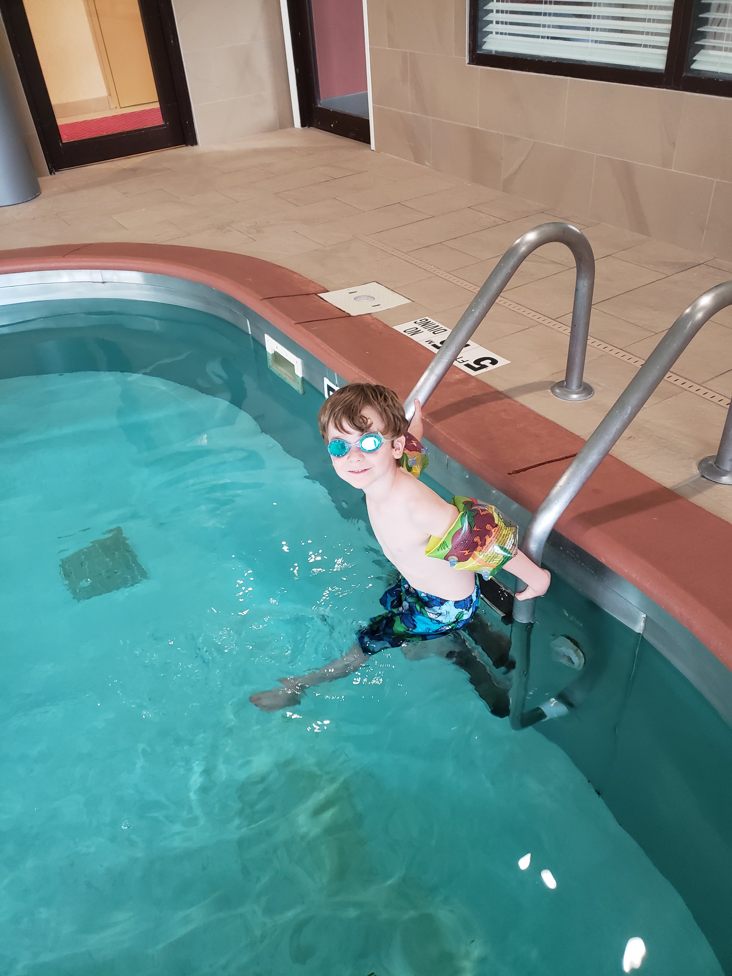 a boy stepping off a pool ladder wearing swim goggles and arm floaties