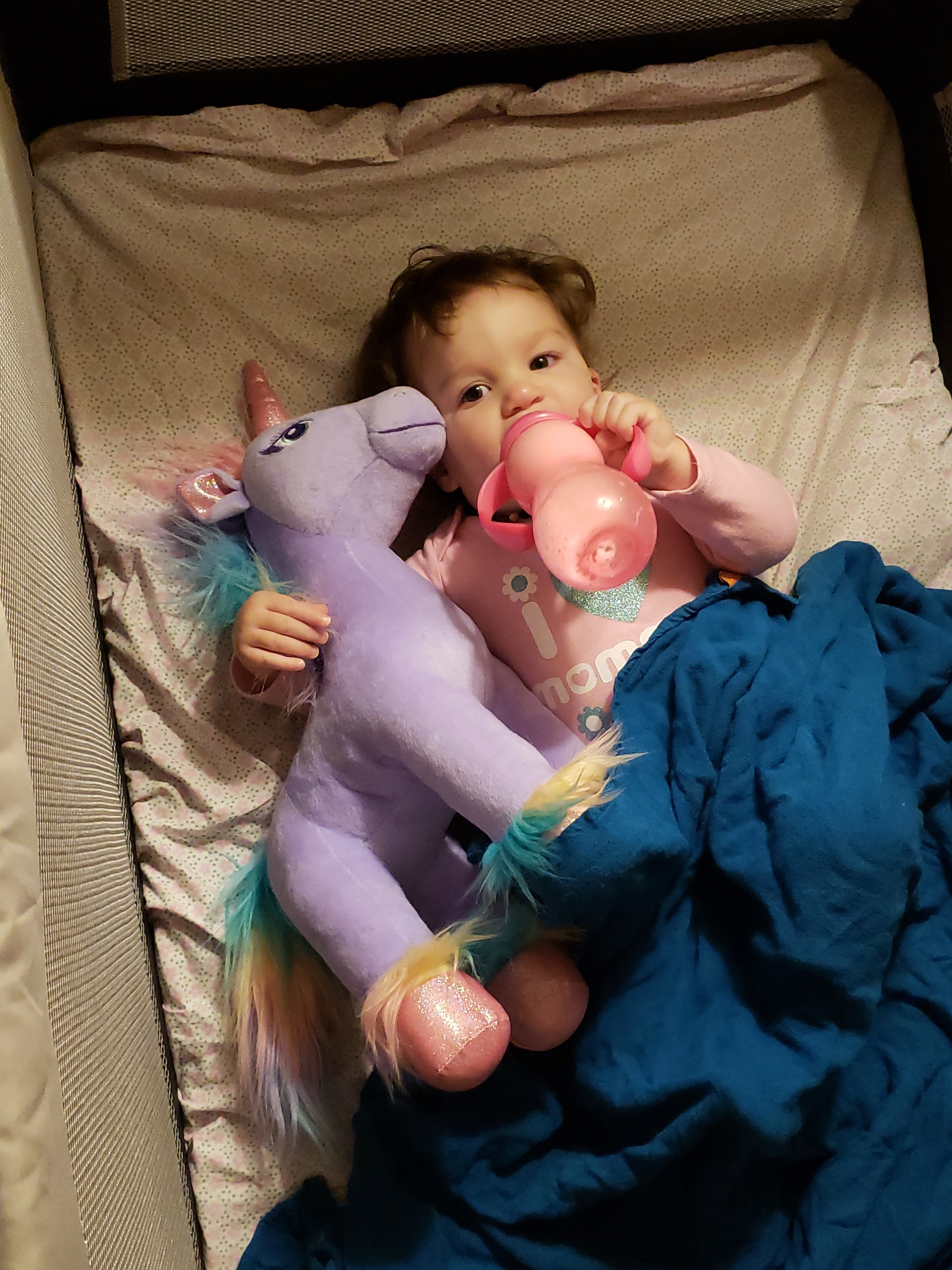 toddler girl in a pack and play with a sippy cup of milk and stuffed unicorn