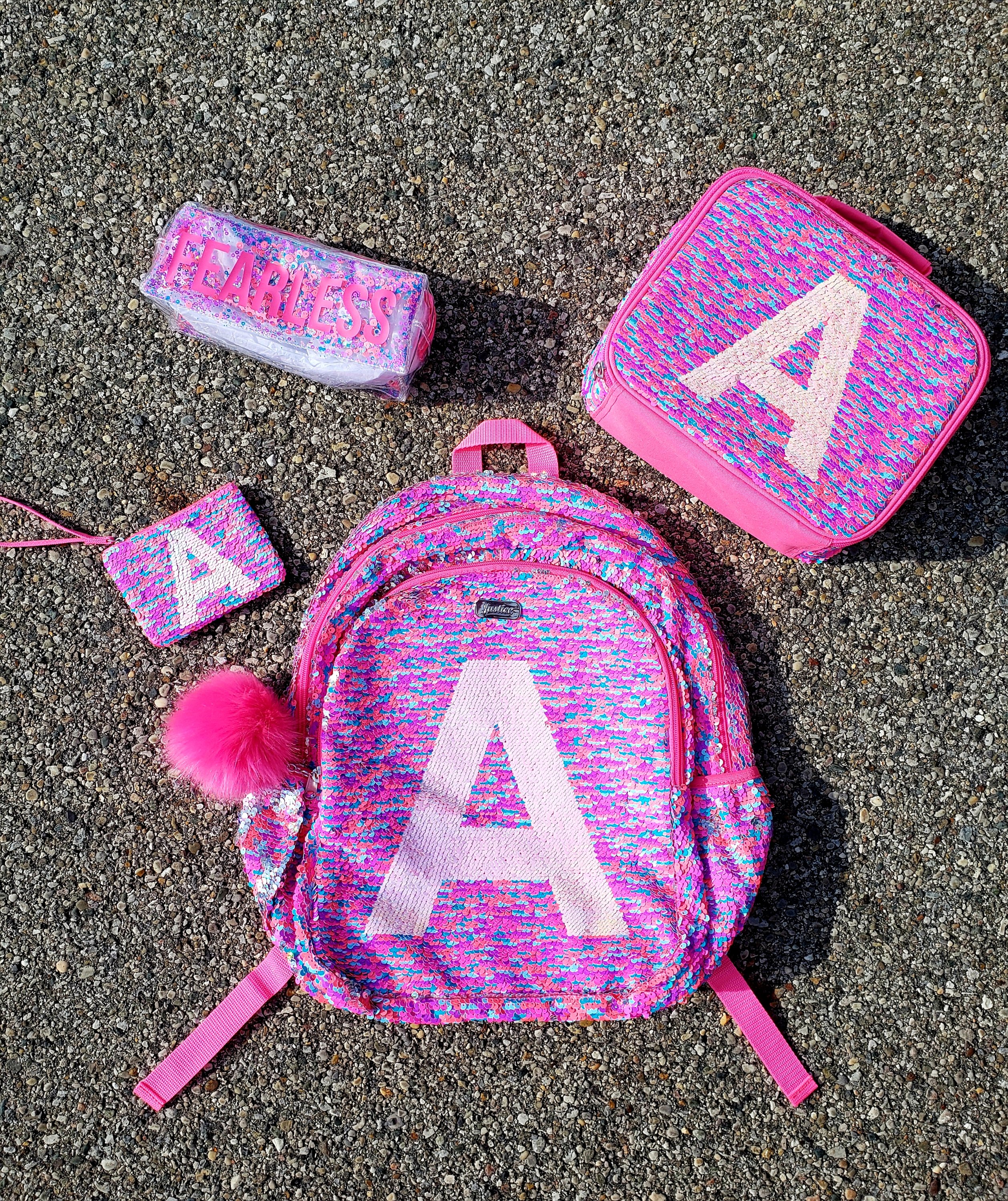 Pink sparkle backpack, lunch kit, coin purse with an A on them, and a "Fearless" pencil case laying on the pavement