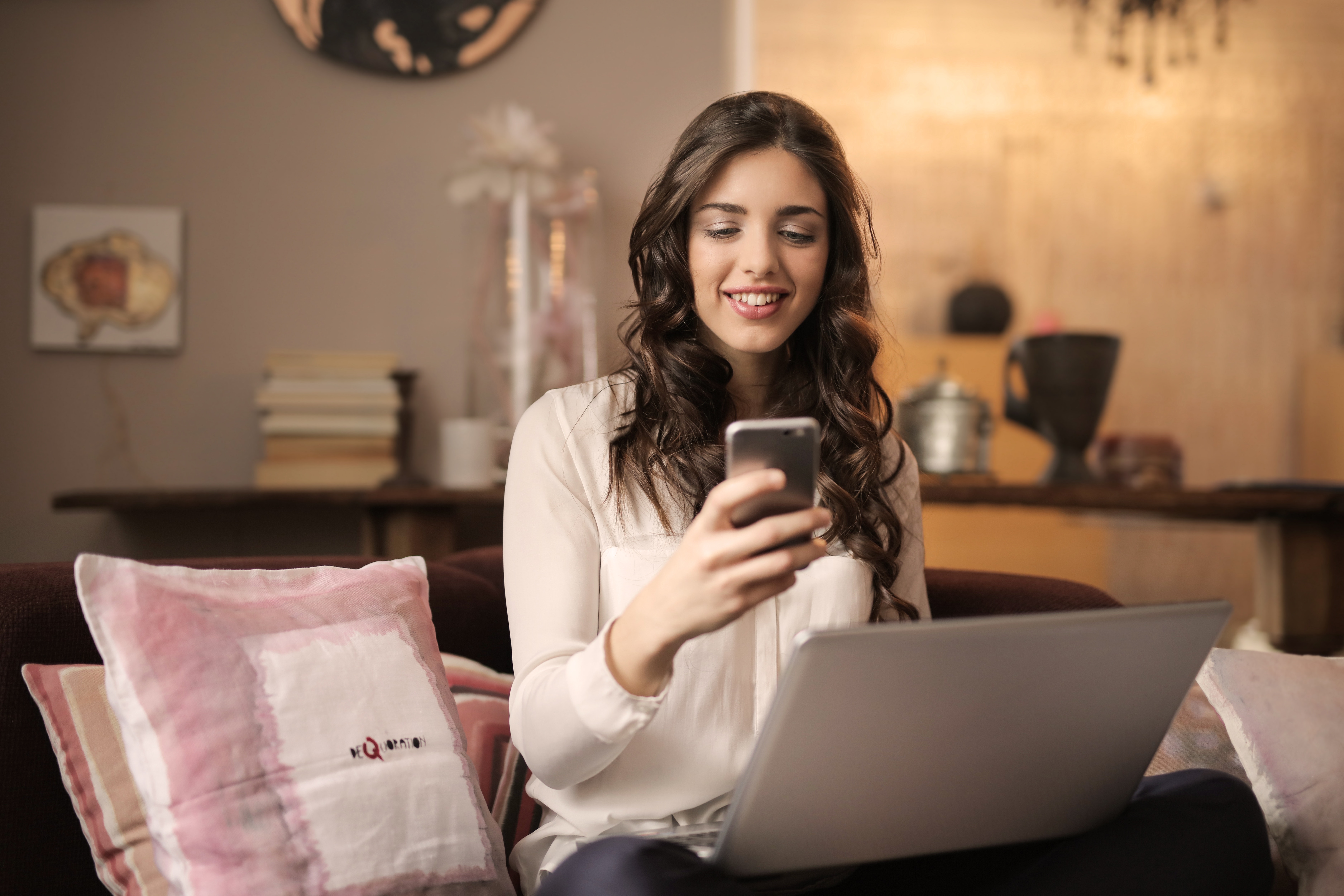 woman sitting on a sofa wit laptop on her lap while looking at her cellphone