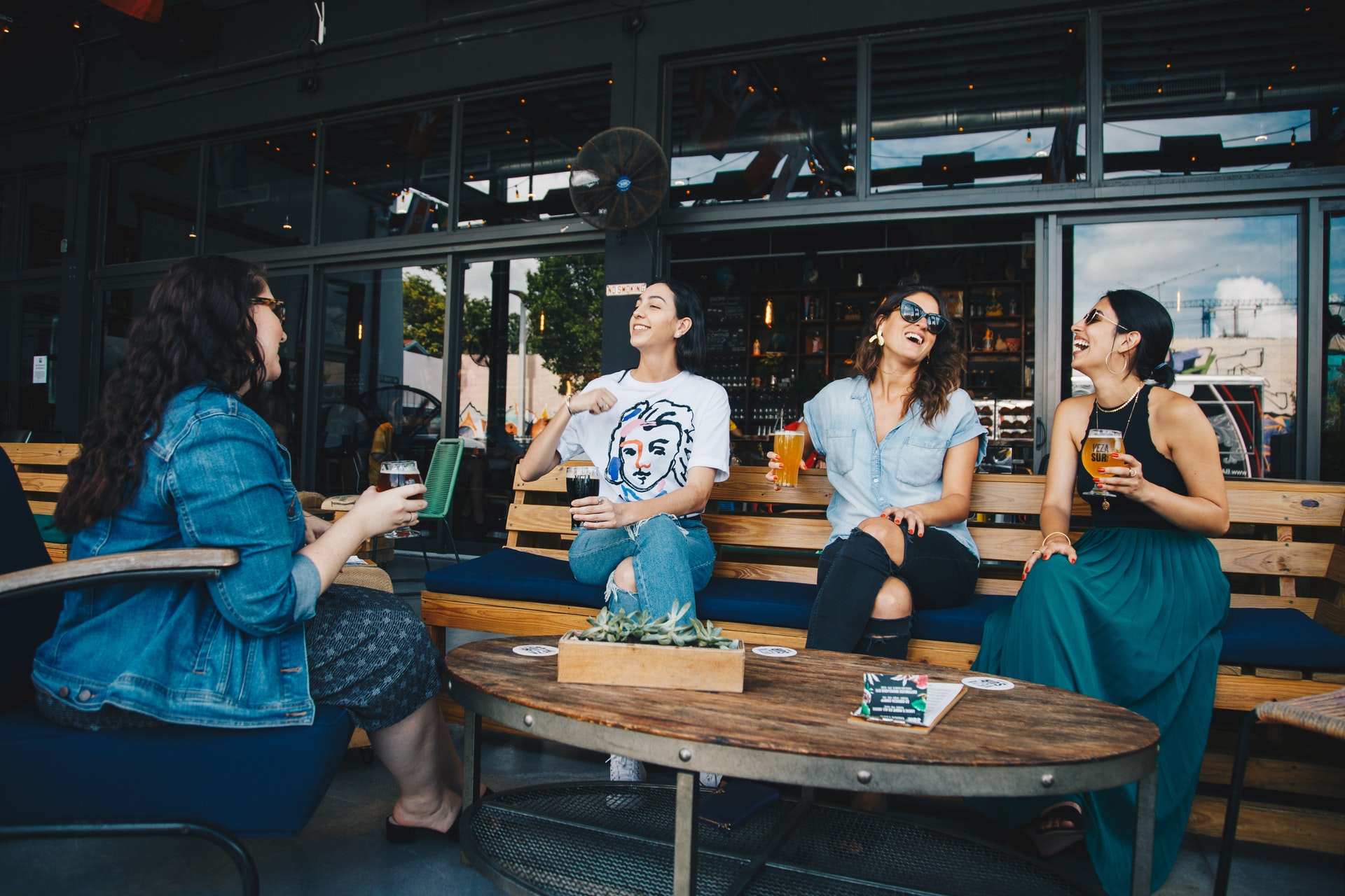 4 women sitting outside on a benches laughing and holding drinks