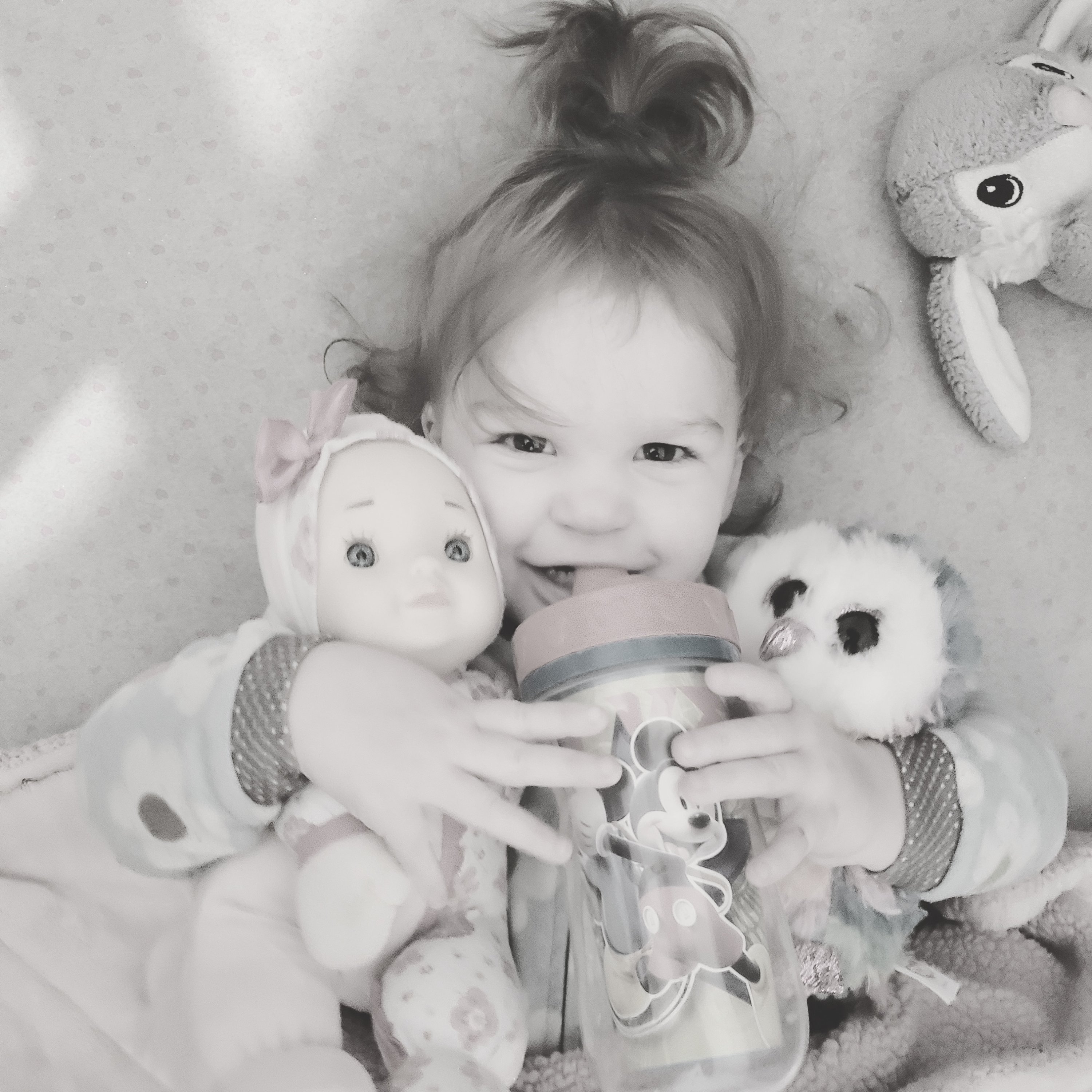 toddler girl laying in bed with a toy in each arm, drinking from a sippy cup, photo in black and white