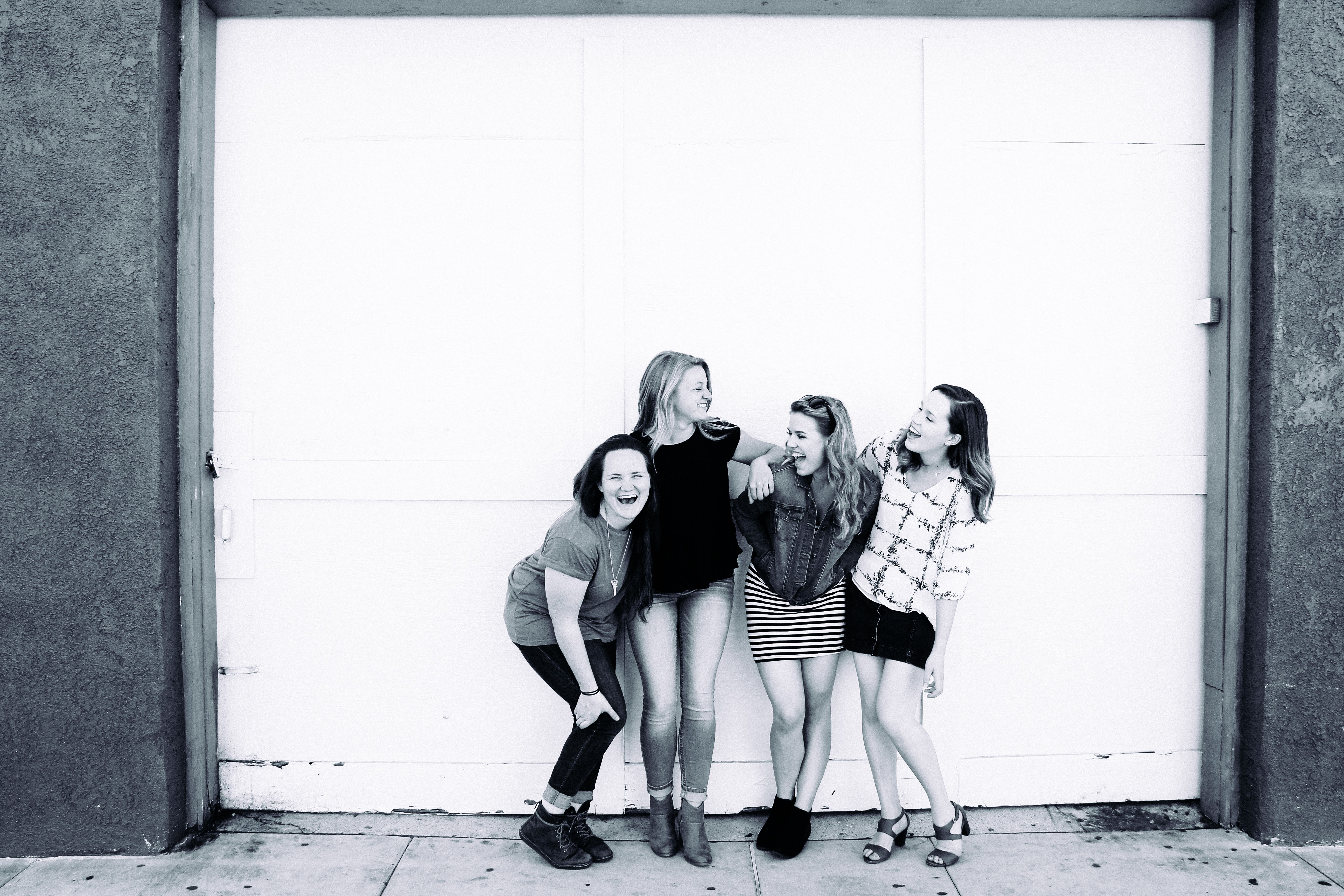 4 women standing together and laughing in front of a white garage door, photo is in black and white