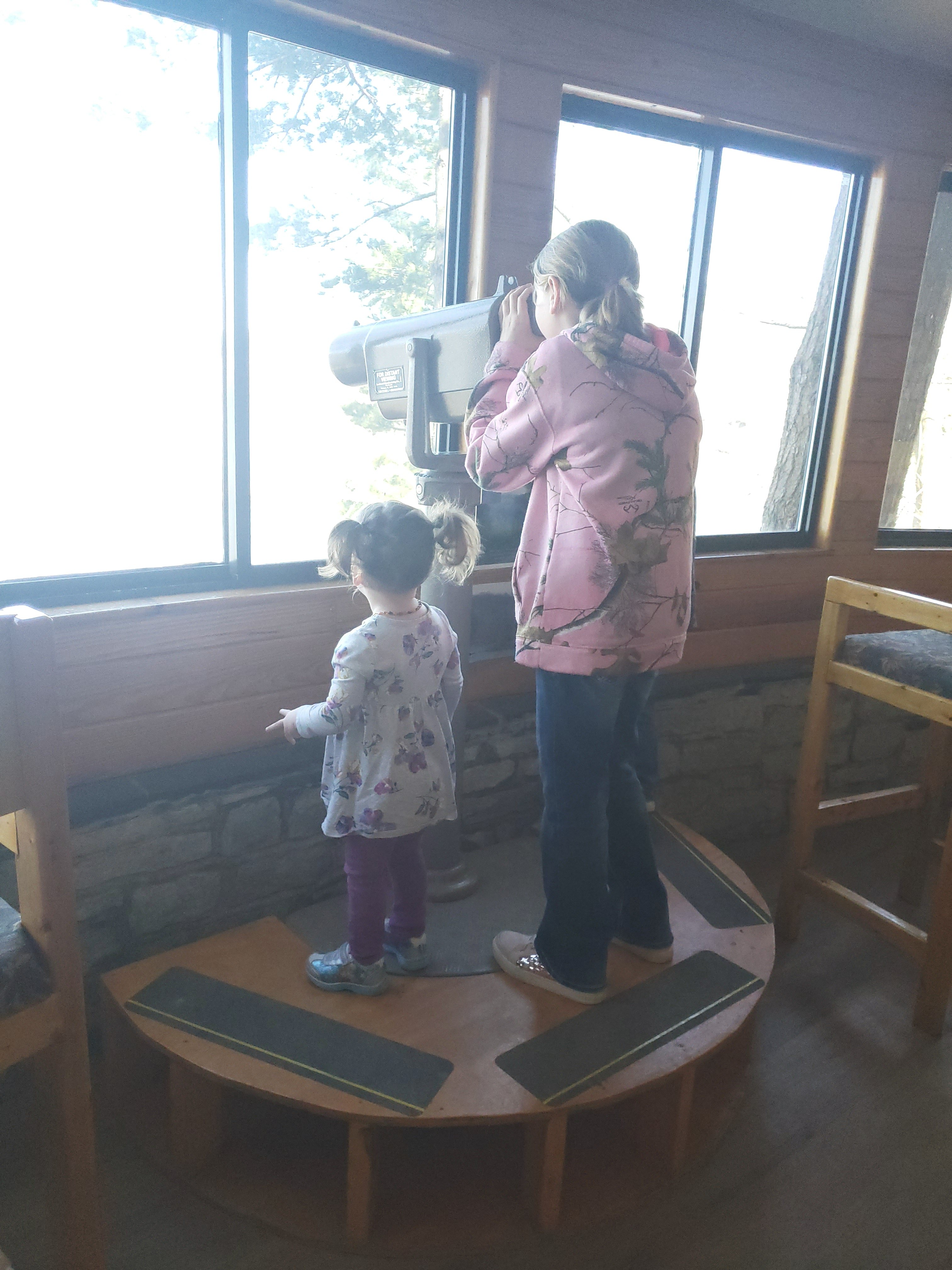 preteen girl looking out a window through a telescope with a toddler standing beside her