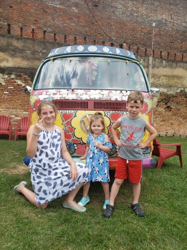 three different aged children standing in front of an old, painted Volkswagen bus