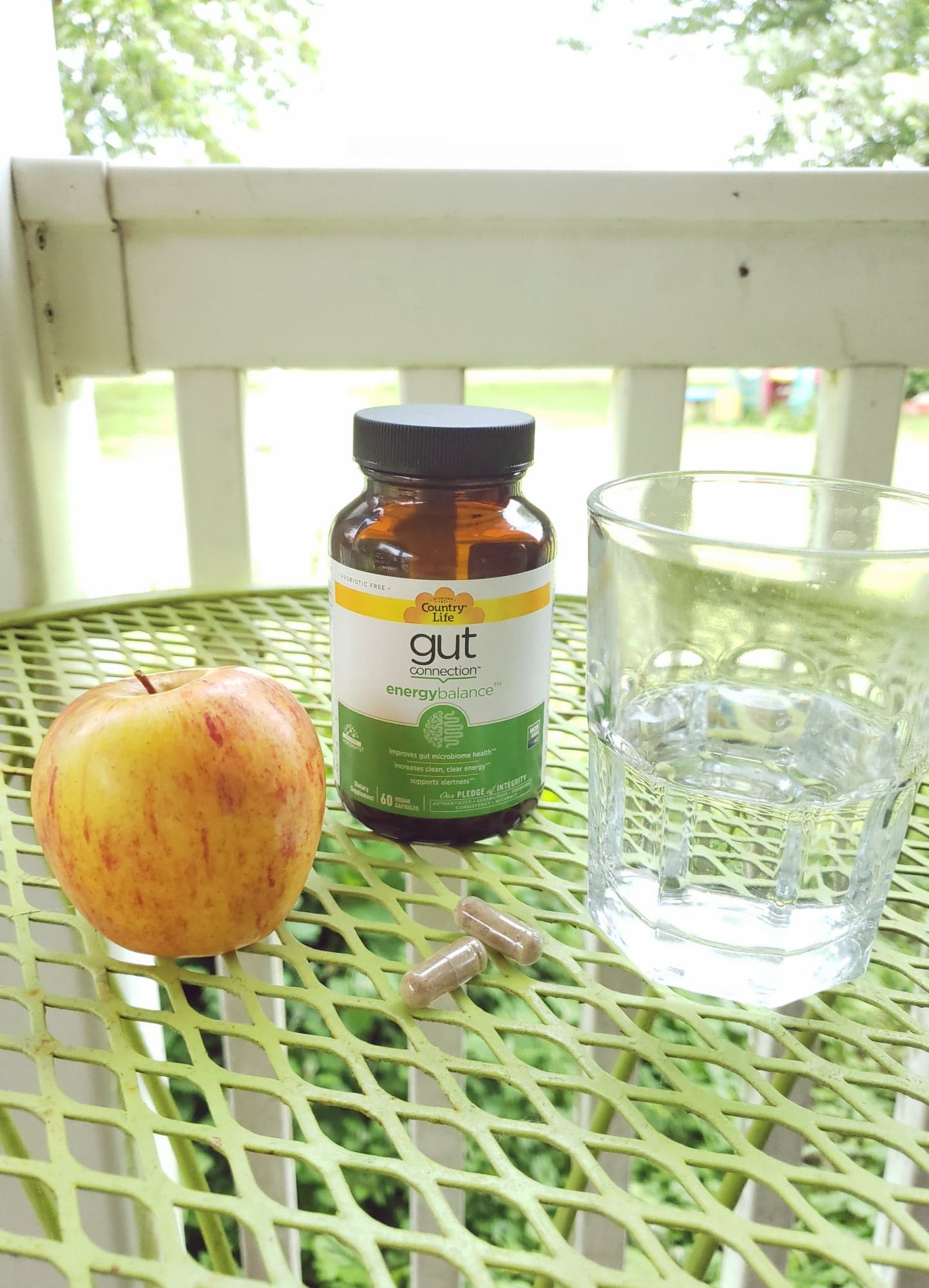 an apple, a glass of water and a bottle of Gut Health Energy Balance pills on a patio table