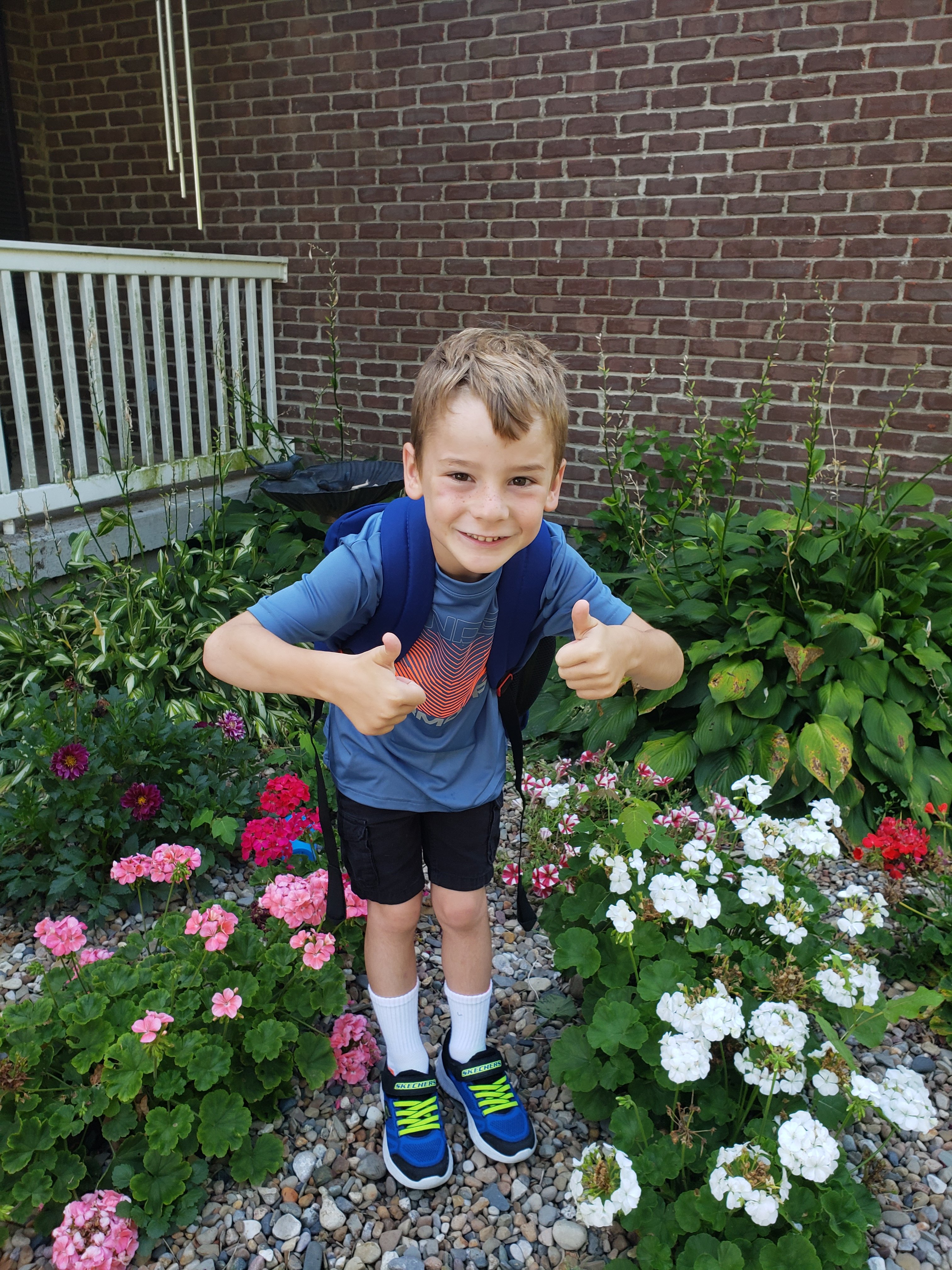 boy standing in flowers in front of a brick wall wearing a backpack giving two thumbs up