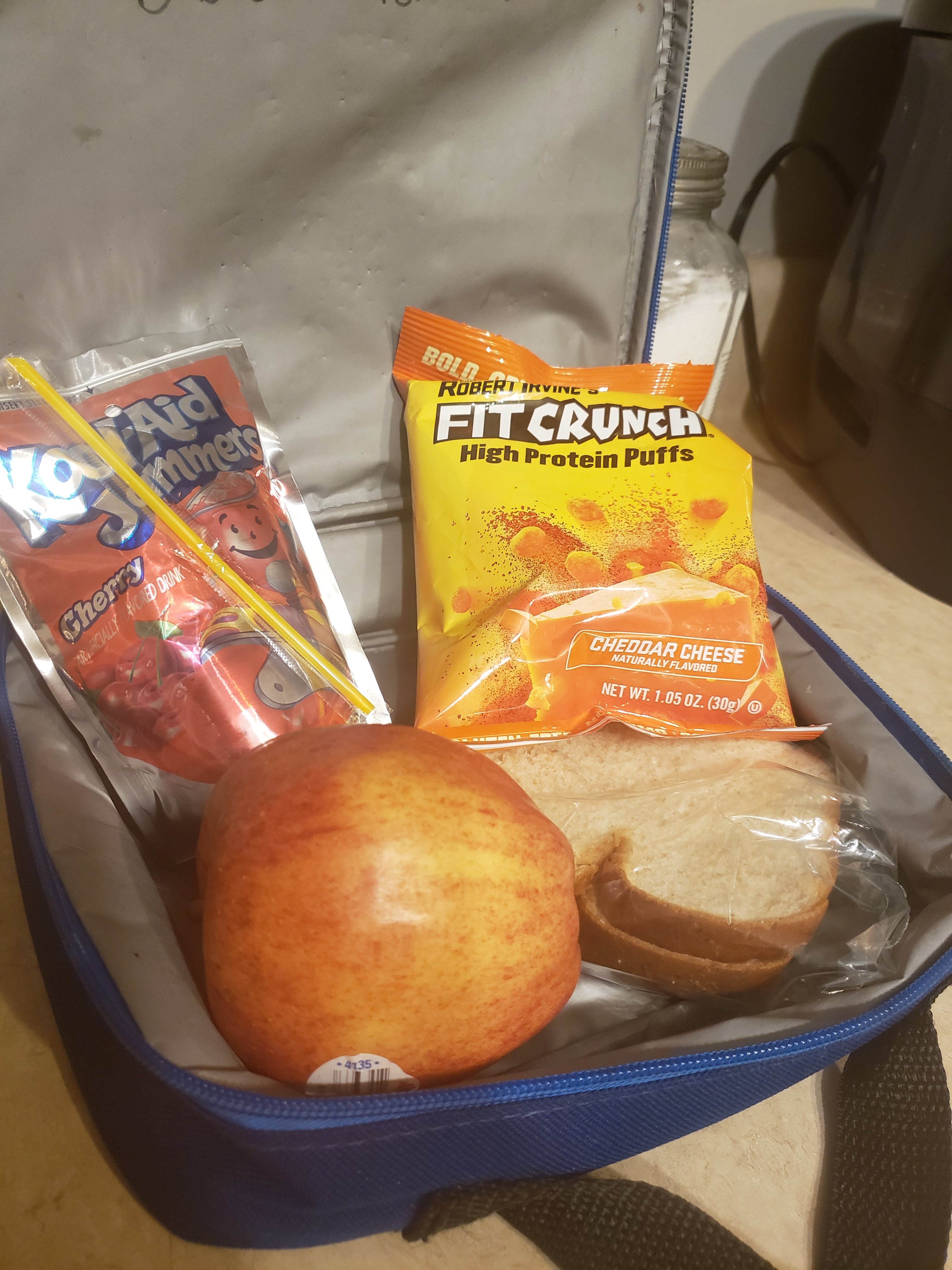 FitCrunch High Protein Puffs in a lunch bag with an apple, a sandwich, and a Kool-Aid Jammers